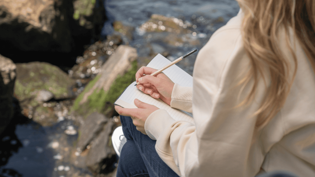 A woman pensively writing in a journal, seated by a serene riverside. | Mind Boost Books - Boost Brain Power with Puzzle Books & Journals