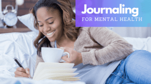 Young woman smiling while journaling in bed with a cup of coffee, depicting the therapeutic practice of journaling for mental health.